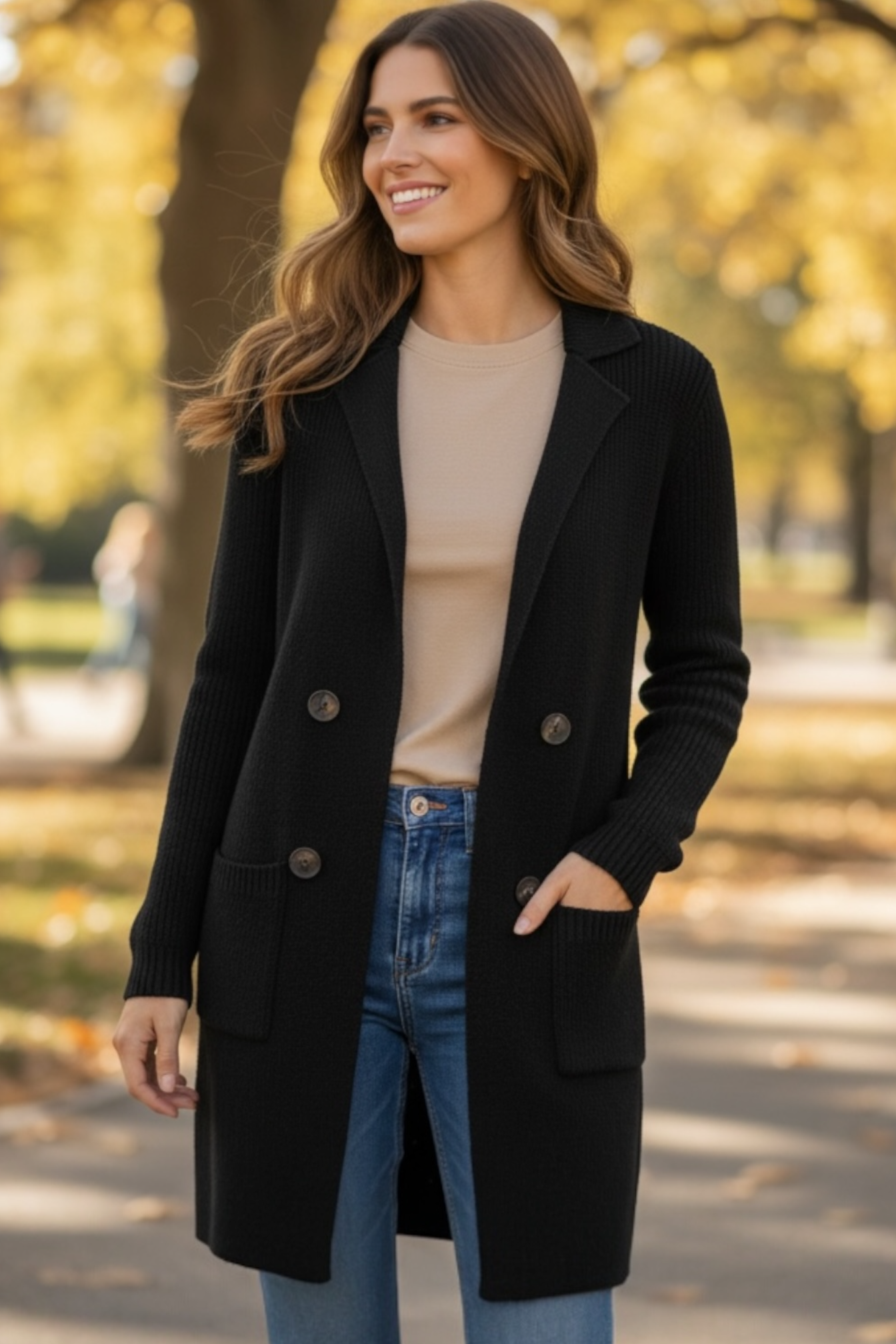 Woman wearing a black coat, beige sweater, and blue jeans standing in an outdoor setting with trees and blurred people in the background.