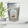 Packaged mud mask powder with a plant in the background on a white surface