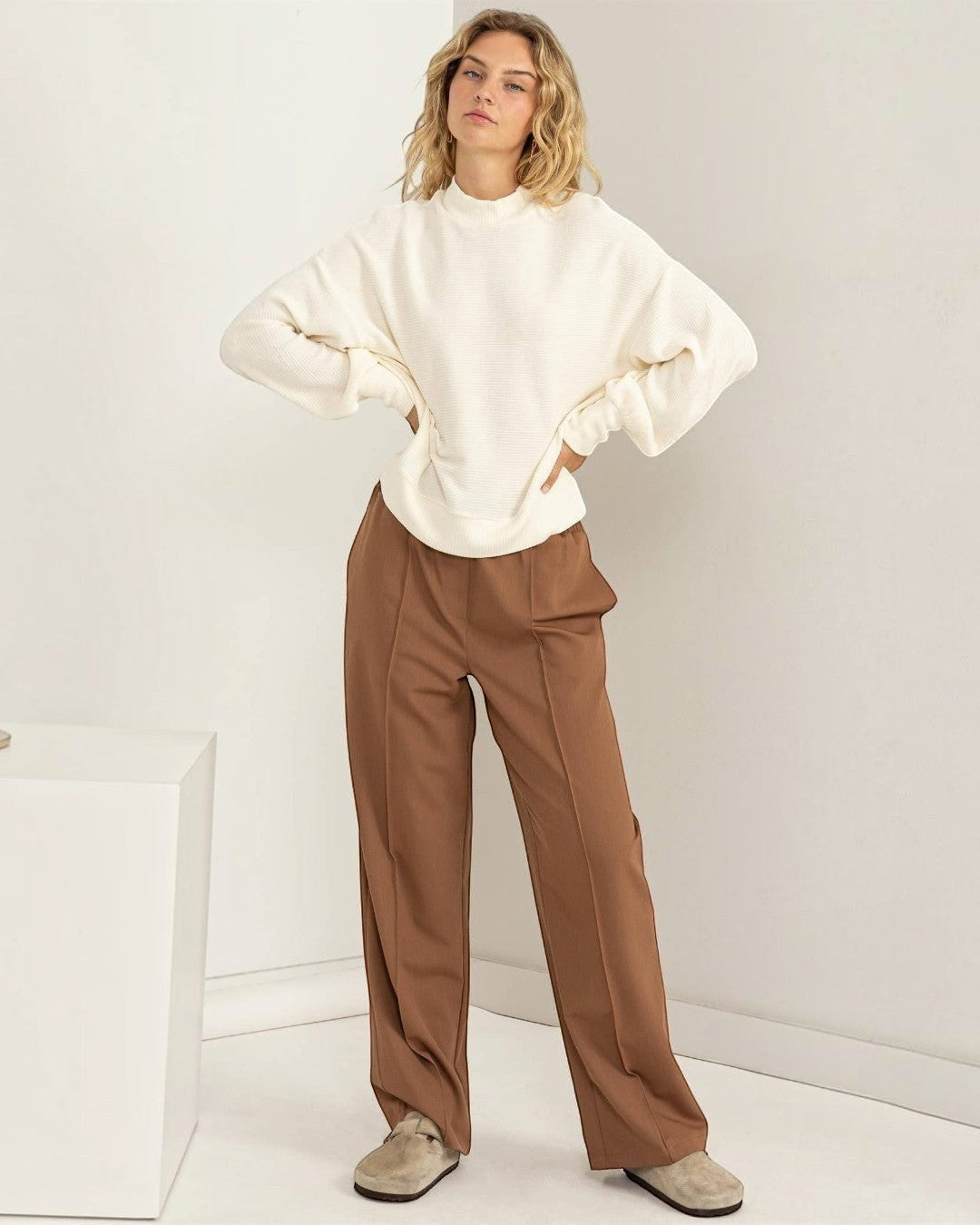 Woman wearing a white sweater and brown pants against a white background