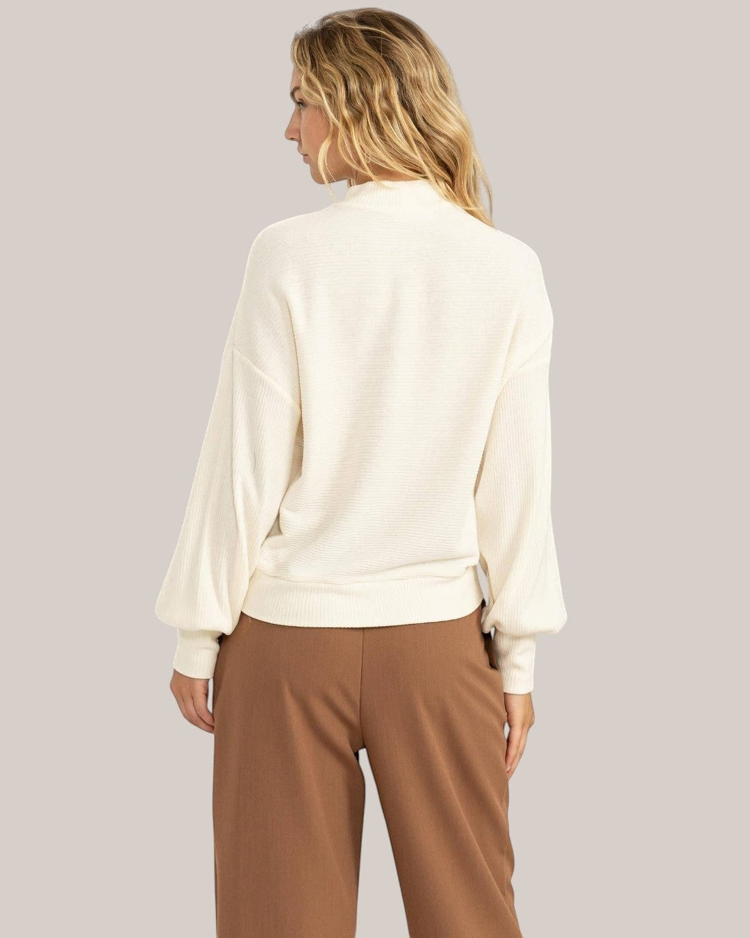 Woman wearing a white blouse and brown pants against a plain background
