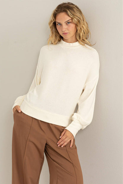 Woman wearing a white sweater and brown pants against a plain background