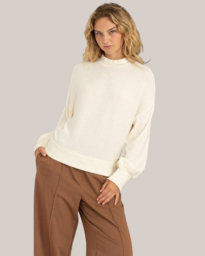 Woman wearing a white sweater and brown pants against a plain background