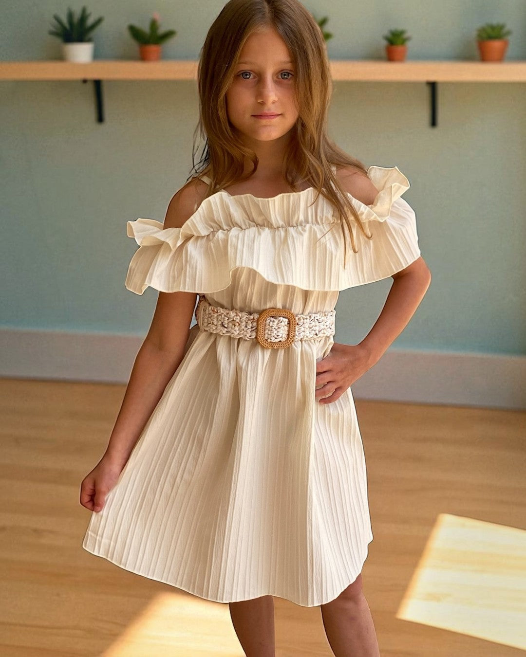 Young girl wearing a beige off-shoulder dress with a belt, standing indoors.