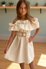 Young girl wearing a beige off-shoulder dress with a belt, standing indoors.