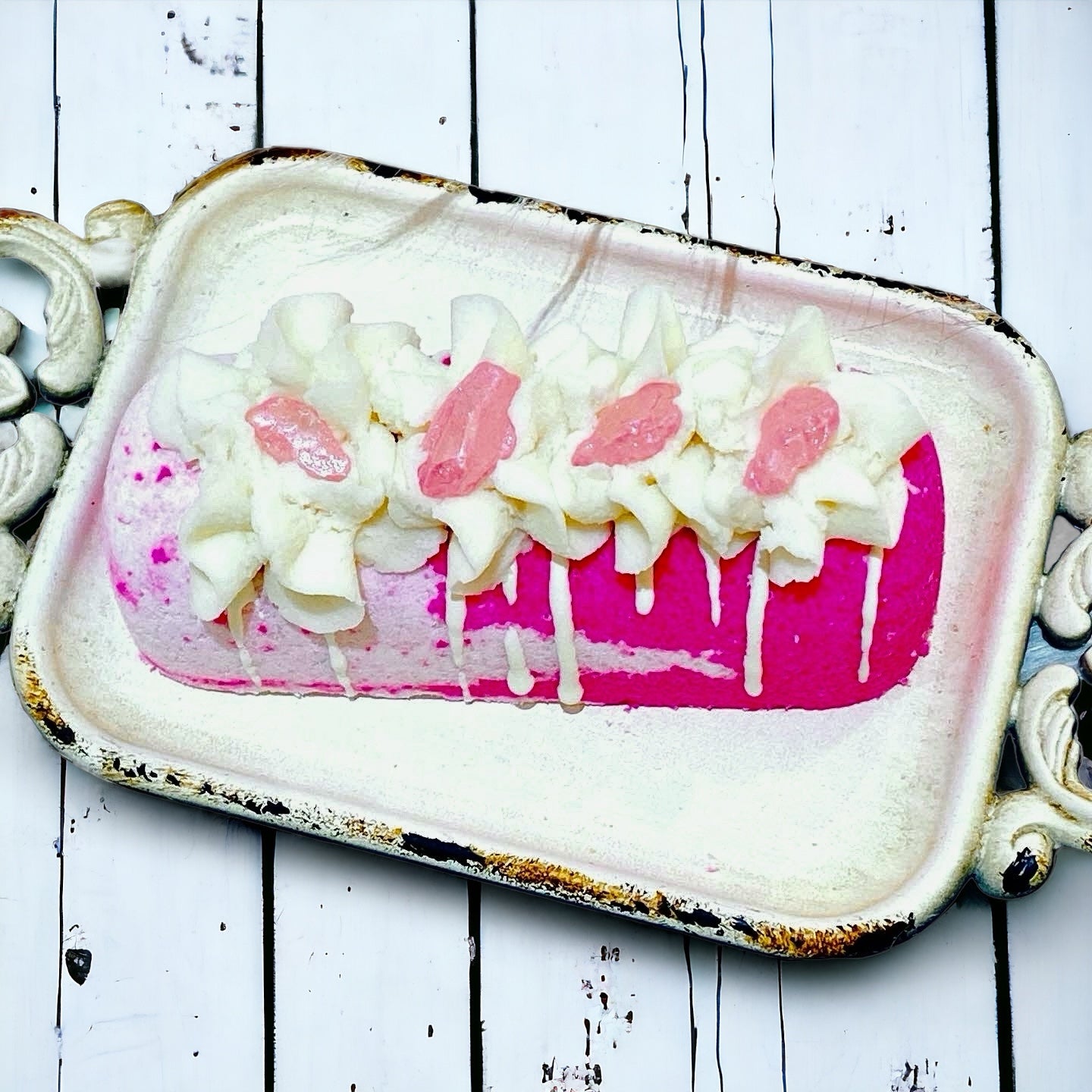 A pink and white twinkie bath bomb topped with white frosting and pink sugar sprinkles on a white tray against a wood background