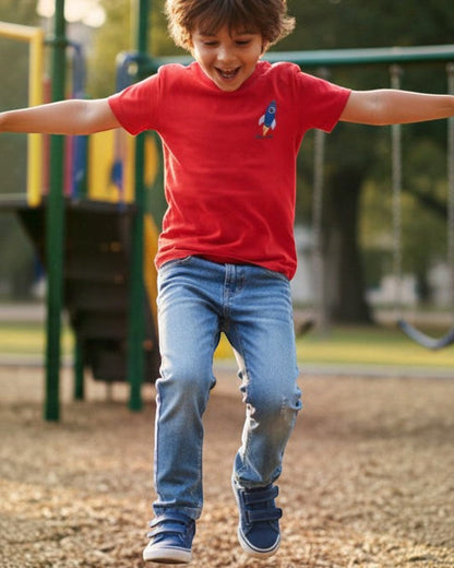 Child in a red shirt and blue jeans jumping on a playground with a slide and swings in the background.
