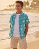 Person wearing a blue floral shirt and beige shorts standing on a beach with palm trees in the background.