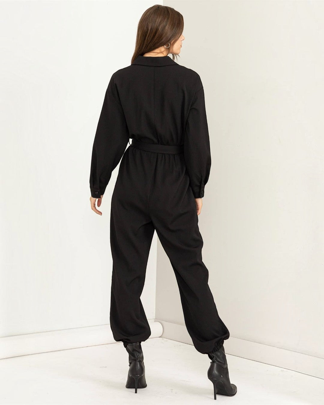 Person wearing a black jumpsuit standing against a white wall.