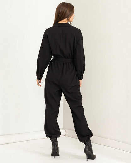 Person wearing a black jumpsuit standing against a white wall.