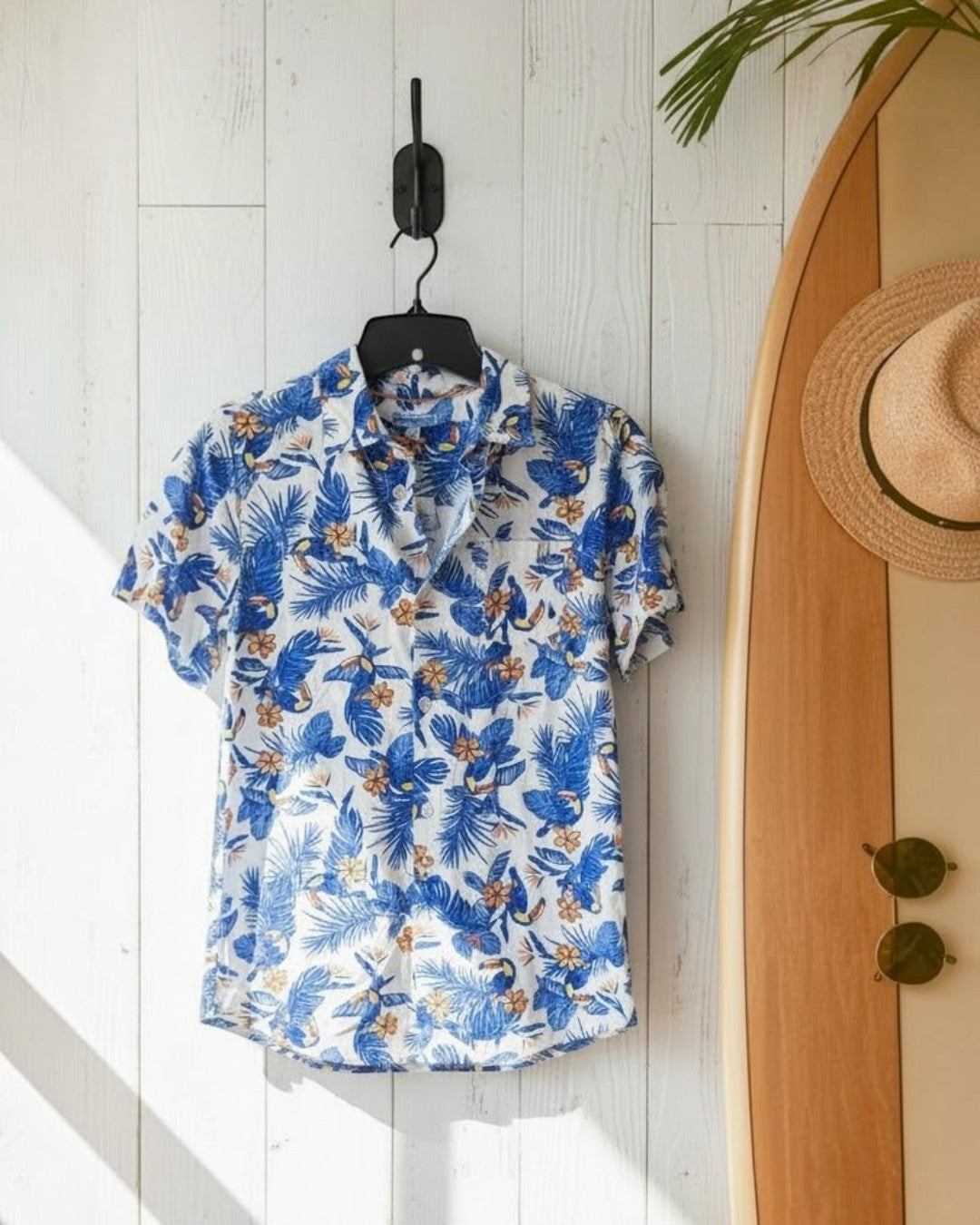 Blue floral shirt on a hanger against a white wooden wall with a surfboard in the background.