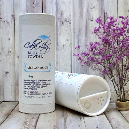 Body powder container labeled 'Calla Lily' with 'Grape Soda' scent on a wooden surface with purple flowers.