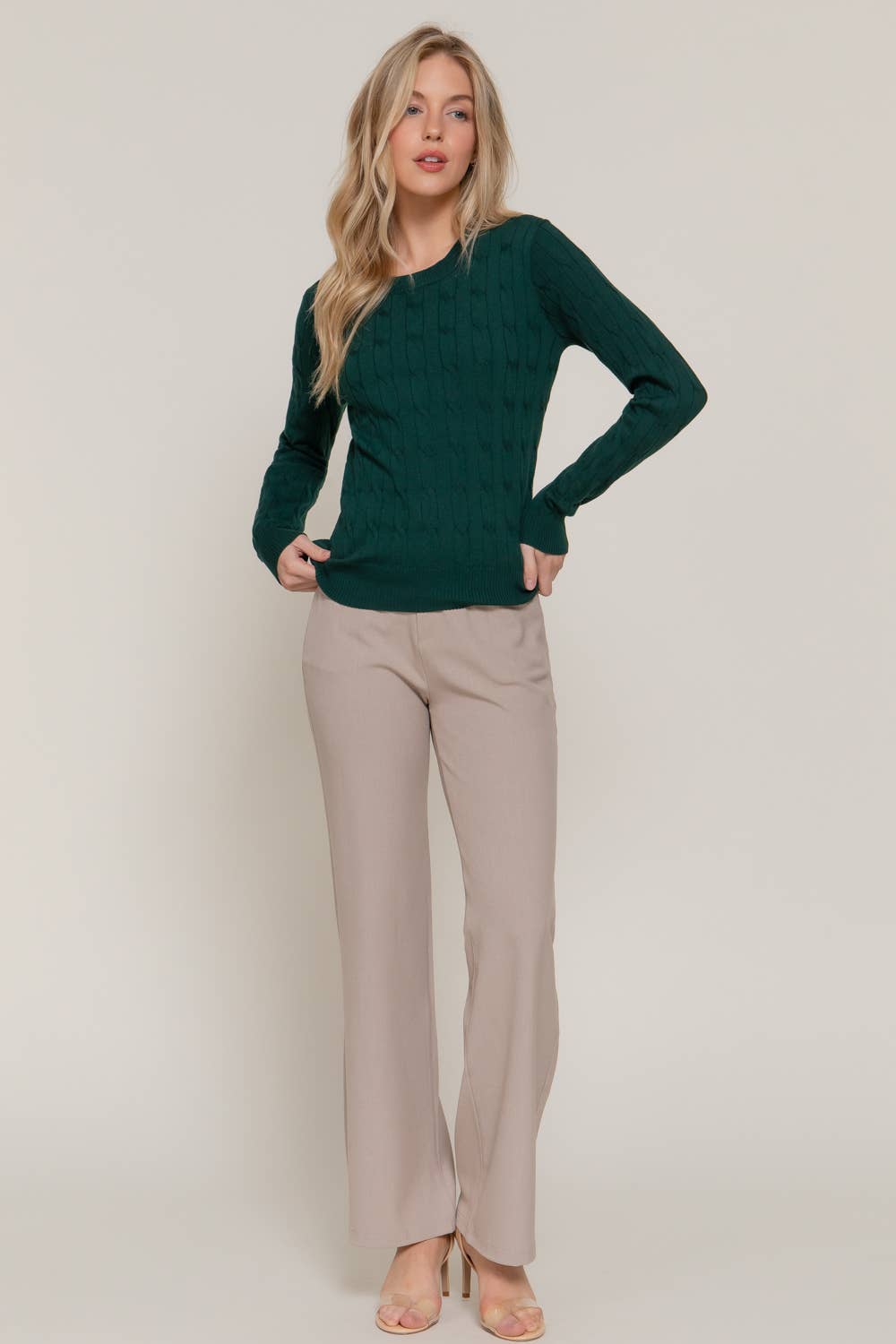 Woman wearing a green sweater and beige pants against a plain background