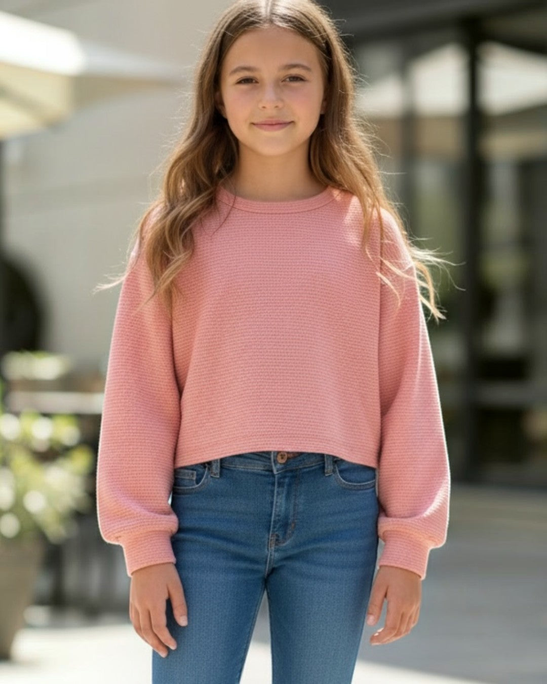 Young girl wearing a pink sweater and blue jeans standing outdoors.