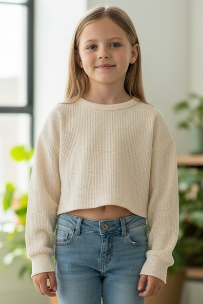 Young girl wearing a beige sweater and blue jeans standing in a bright room with plants.