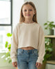 Young girl wearing a beige sweater and blue jeans standing in a bright room with plants.