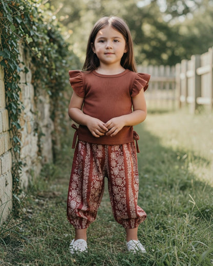 Young girl standing outdoors in a natural setting wearing a rust-colored top and patterned pants.