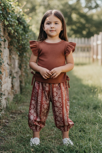 Young girl standing outdoors in a natural setting wearing a rust-colored top and patterned pants.