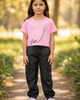 Young girl wearing a pink shirt with a design and text, black cargo pants, and white sneakers standing on a path in a park.