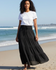 Woman wearing a white t-shirt and black long skirt standing on a beach with ocean waves in the background.