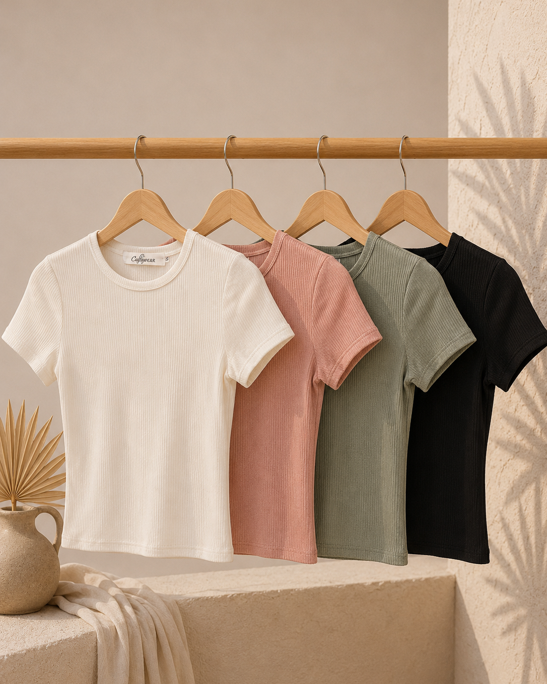 Four t-shirts in white, pink, green, and black hanging on a rack against a neutral background.
