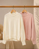 Two sweaters, one cream and one pink, hanging on a wooden rack against a neutral background.