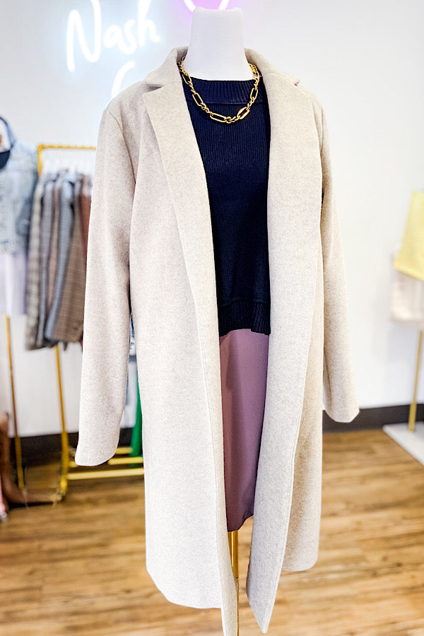 Mannequin wearing a long beige cardigan over a dark top and purple skirt in a store setting.
