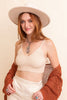 Woman wearing a beige top, white pants, and a rust-colored cardigan with a beige hat on a beige background.