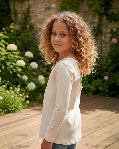 Young girl with curly hair wearing a white long-sleeve shirt in a garden setting.