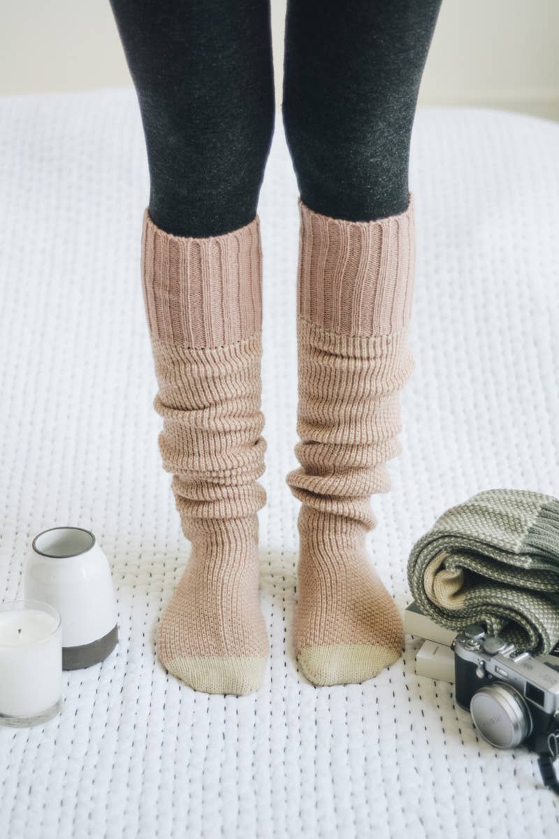 Person wearing beige ribbed knee-high socks on a white surface with a camera and folded blanket.