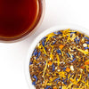 A close-up overhead shot of loose-leaf herbal tea in a white bowl, with a glass of dark amber tea at the top left. The tea blend contains brown tea leaves mixed with bright yellow, blue, and orange dried flower or citrus pieces on a white background.