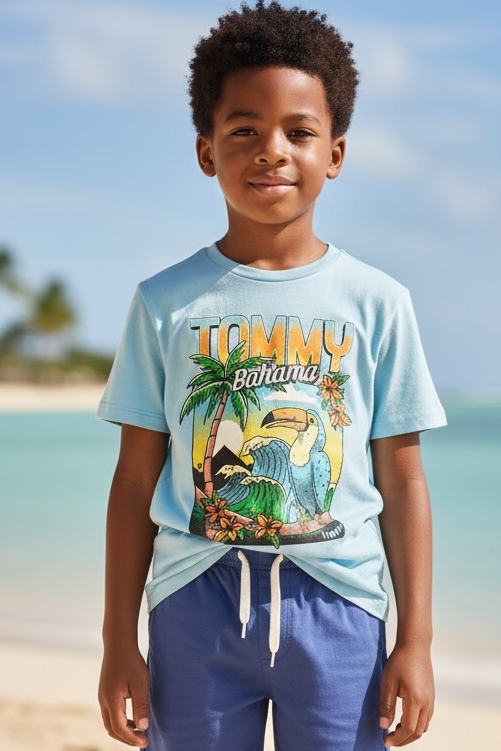 Child wearing a light blue t-shirt with a graphic design and blue shorts standing on a sandy beach with ocean and sky in the background.