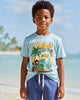 Child wearing a light blue t-shirt with a graphic design and blue shorts standing on a sandy beach with ocean and sky in the background.