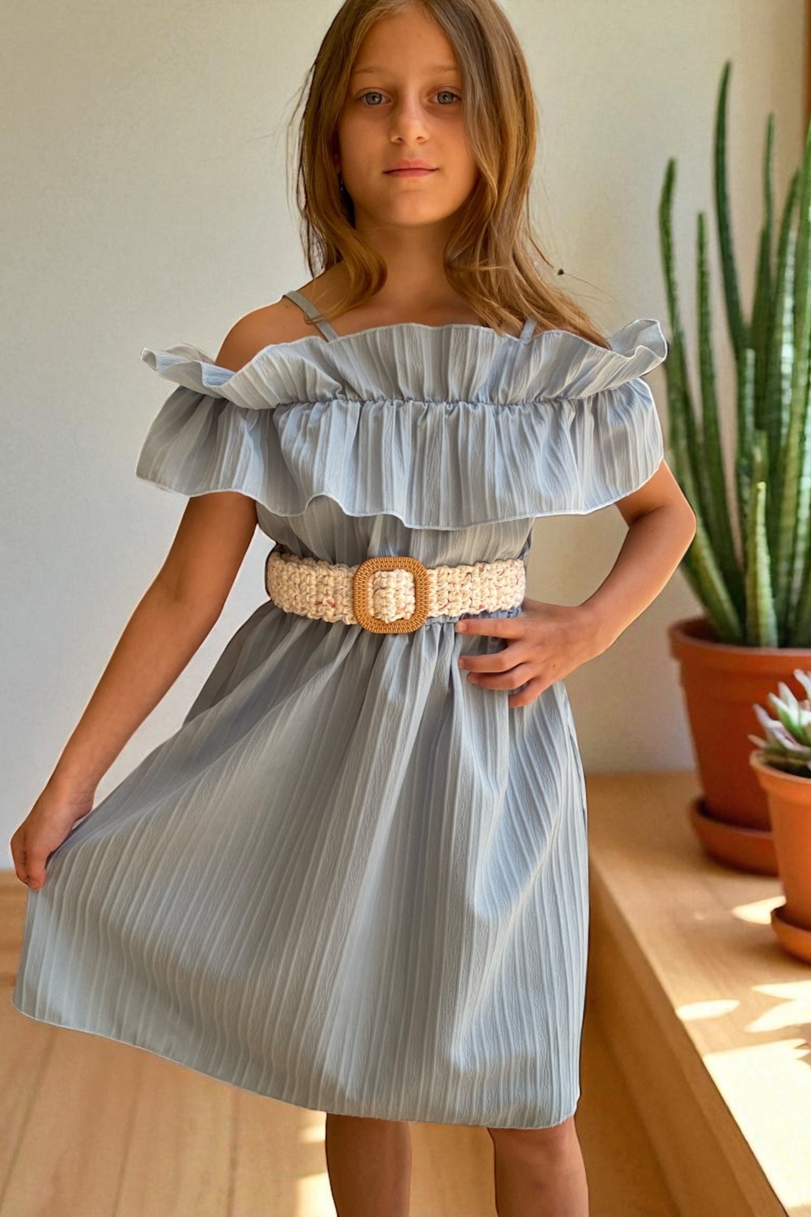Young girl wearing a light blue dress with a belt, standing indoors with plants in the background.