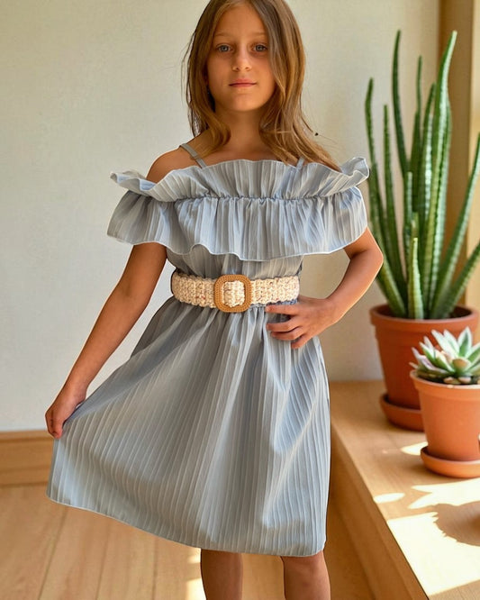 Young girl wearing a light blue dress with a belt, standing indoors with plants in the background.