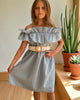 Young girl wearing a light blue dress with a belt, standing indoors with plants in the background.