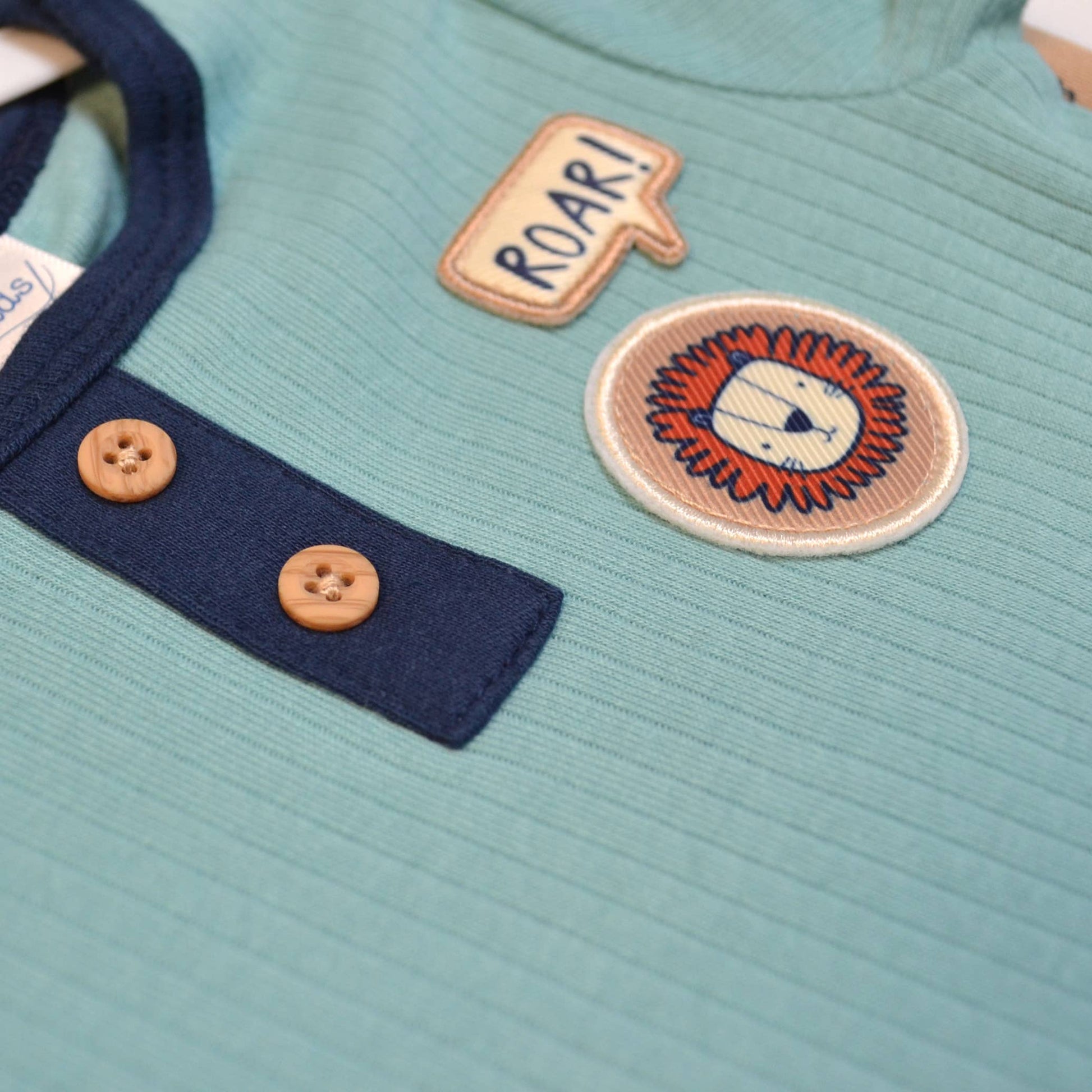 Close-up of a teal garment with a lion patch and 'ROAR!' text, featuring buttons.