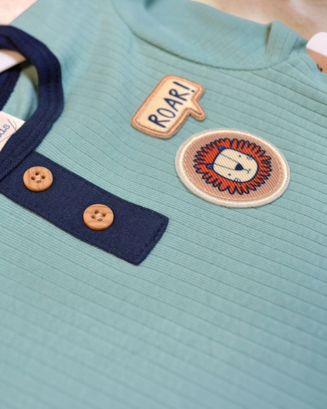 Close-up of a teal garment with a lion patch and 'ROAR!' text, featuring buttons.