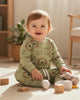 Baby sitting on a light-colored floor with wooden blocks and a plush toy, wearing a green outfit with animal prints.