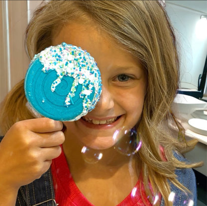 Child holding a blue cookie with sprinkles up to their eye, smiling.