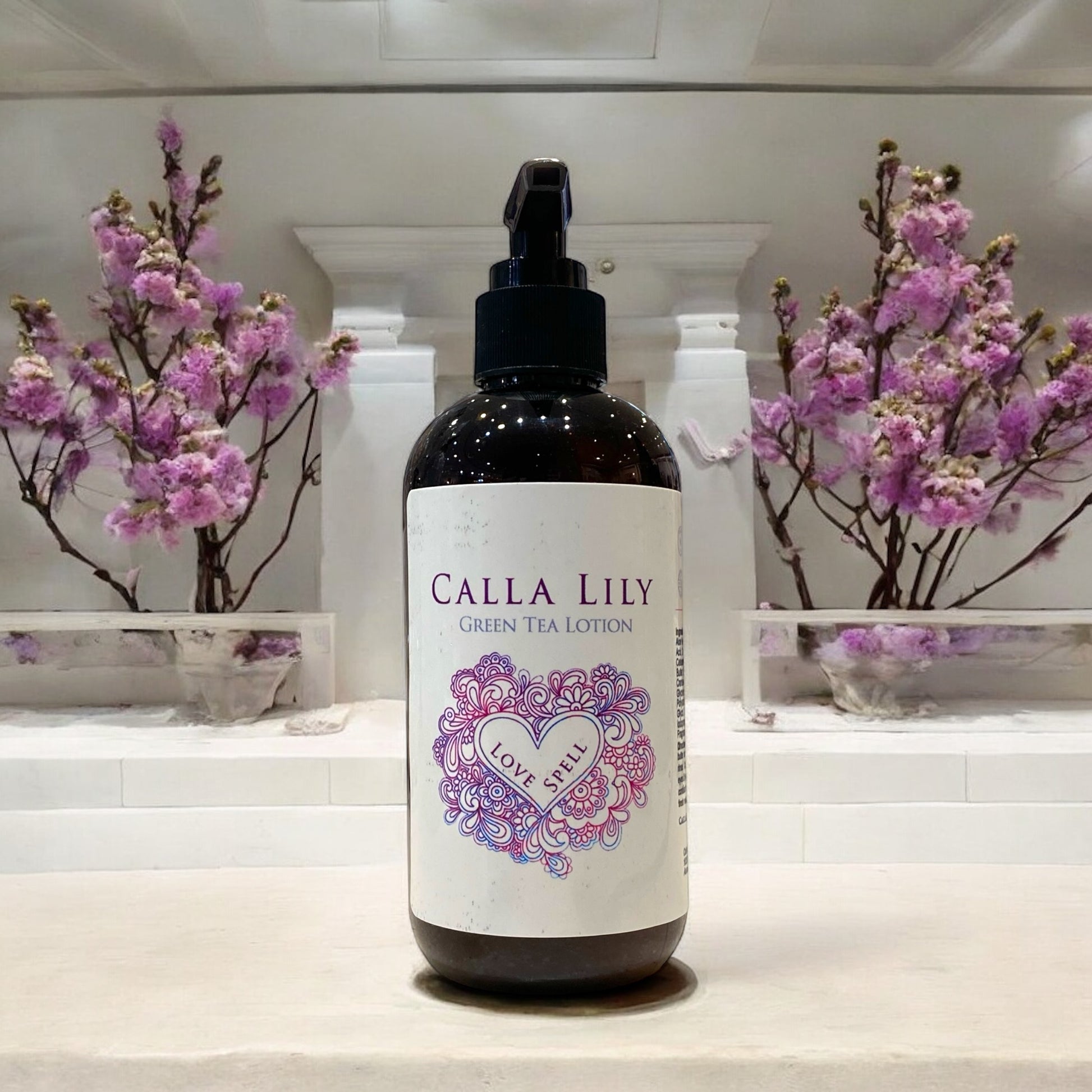 Bottle of Calla Lily Green Tea Lotion with decorative label on a white surface with pink flowers in the background.