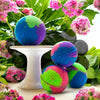 Colorful bath bombs on a white stand with pink flowers and green leaves in the background
