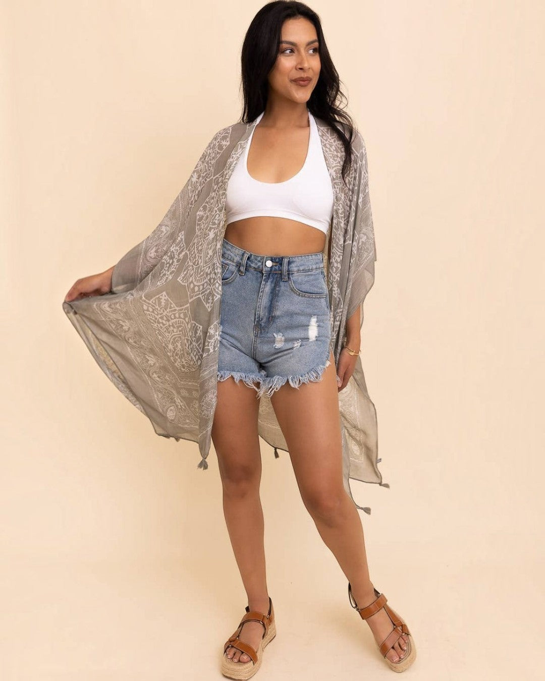 Woman wearing a white crop top, denim shorts, and a long, light-colored kimono against a beige background.