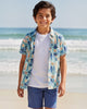 Young boy standing on a beach wearing a colorful shirt and blue shorts.