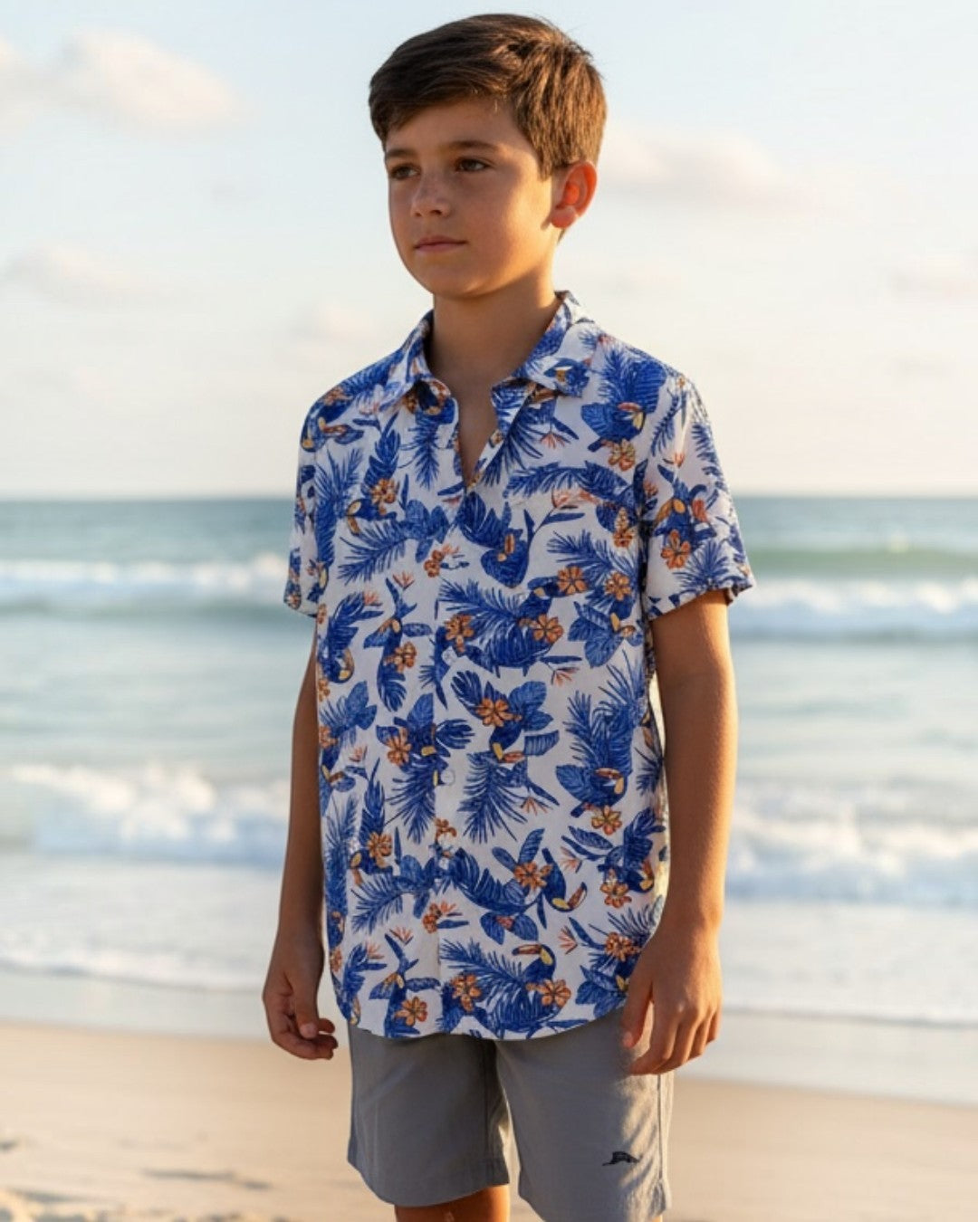 Young boy standing on a sandy beach wearing a blue floral shirt and light gray shorts.