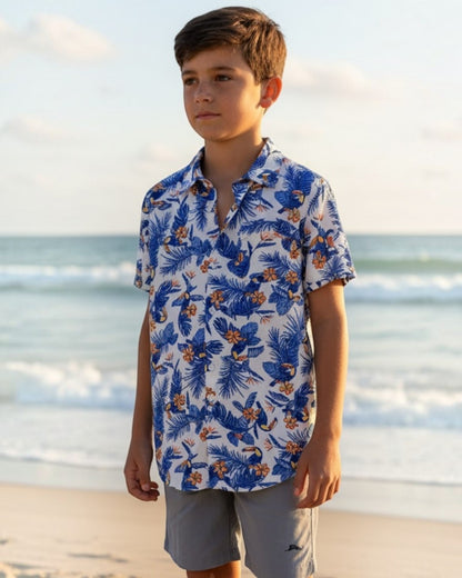 Young boy standing on a sandy beach wearing a blue floral shirt and light gray shorts.