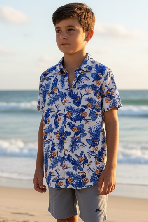 Young boy standing on a sandy beach wearing a blue floral shirt and light gray shorts.
