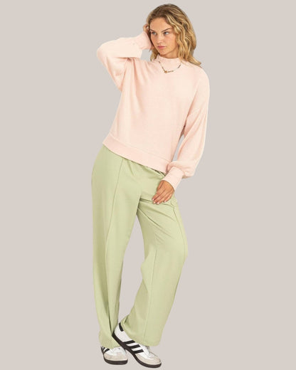 Woman wearing a light pink sweater and green pants against a plain background