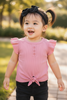Child wearing a pink shirt with ruffled sleeves and a black bow headband outdoors.