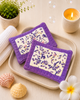 Two purple coasters with white designs on a beige tray, surrounded by decorative items like a candle, plant, and flowers.