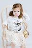 Young girl sitting on a chair wearing a white outfit with a fox design, holding a gold handbag.